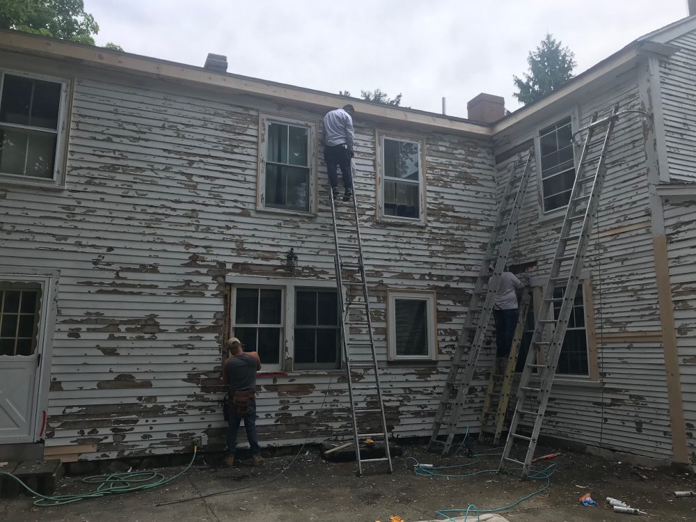 Workmen on ladders stripping paint from the clapboards during the rehabilitation